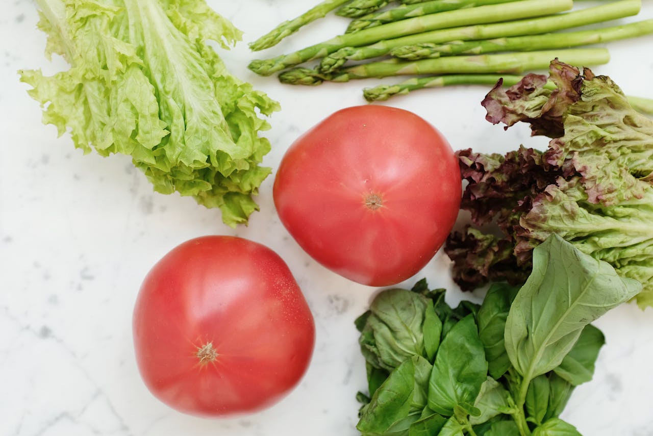 Top view of fresh vegetables including lettuce, asparagus, tomatoes, and basil on marble surface.